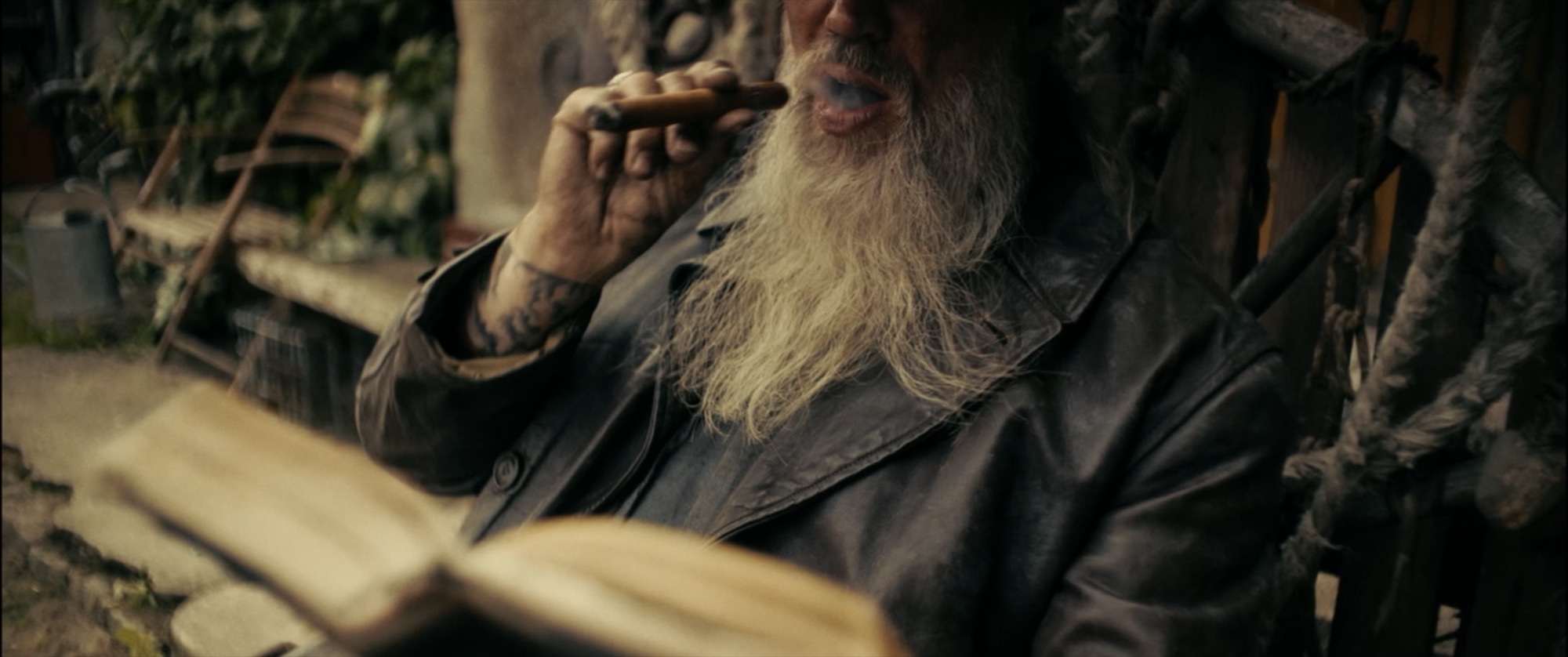 Close-up of a bearded old man in a worn leather coat drawing on a cigar, tattooed hand raised - Amon Amarth
